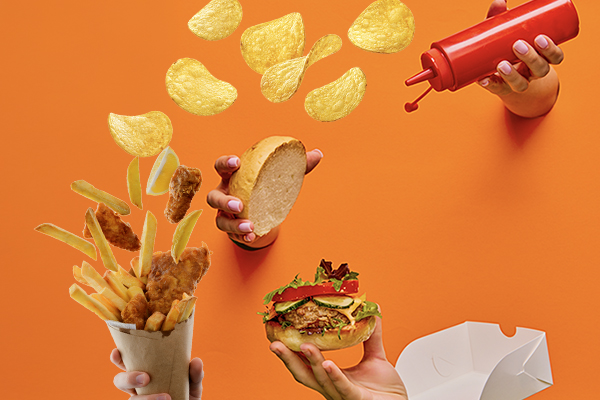Mehrere Hände halten Burger, Ketchup und eine Tüte mit Pommes und Chickennuggets hoch. Im Hintergrund fliegen Kartoffelchips durch das Bild.