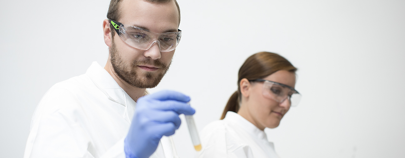 Eine Labormitarbeiterin und ein Labormitarbeiter mit Kittel und Schutzbrille sind zu sehen. Der Labormitarbeiter hält ein Probenröhrchen in der Hand und schaut sich dieses an.